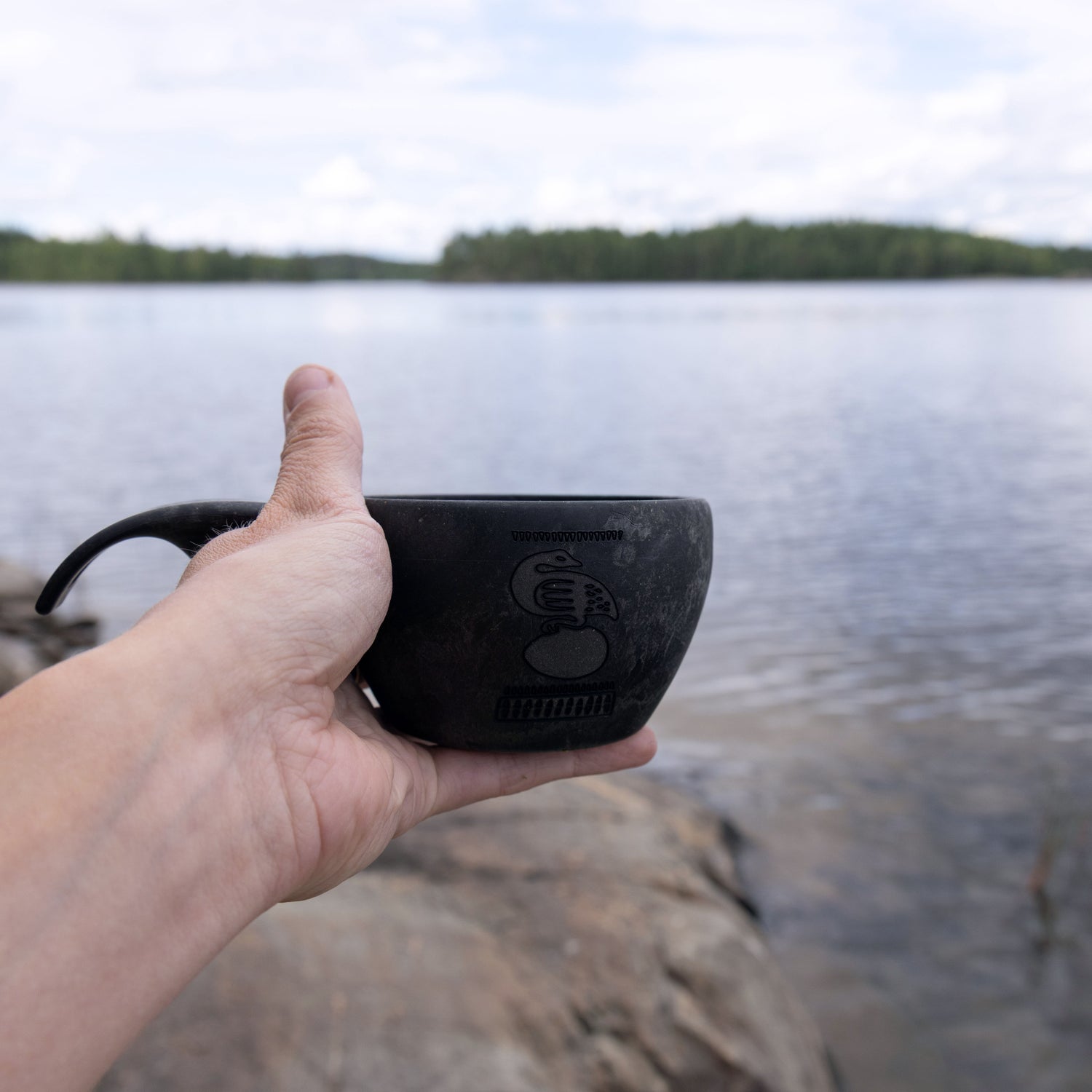 Musta Kalevala Kupilka 21 retkimuki Kalevala Sotka kaiverruksella, taustalla järvimaisema